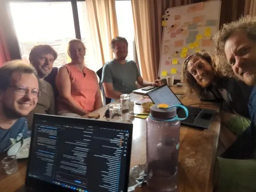 Mehrere Menschen sitzen um einen Tisch mit Laptops, im Hintergrund eine Tafel voller Notizzettel.